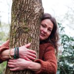 Yoga teacher hugging a tree with a gentle smile, looking directly into the camera