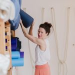 Yoga teacher looking at the camera with a warm smile while reaching for a yoga mat on a shelf.