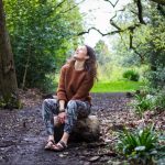 Yoga teacher sitting peacefully on a log, gazing up at a tall tree, surrounded by nature.